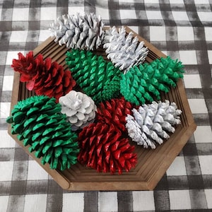 May include: A wooden tray filled with painted pine cones in red, green, and white. The pine cones are arranged on a checkered black and white tablecloth. The tray is octagonal.