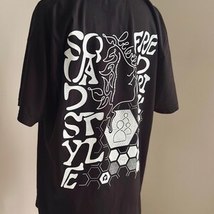 May include: Black t-shirt featuring a white graphic on the back. The design incorporates the words "SQUAD STYLE FREE" in a unique font, a line drawing of a face, and geometric shapes. The t-shirt is displayed on a mannequin.