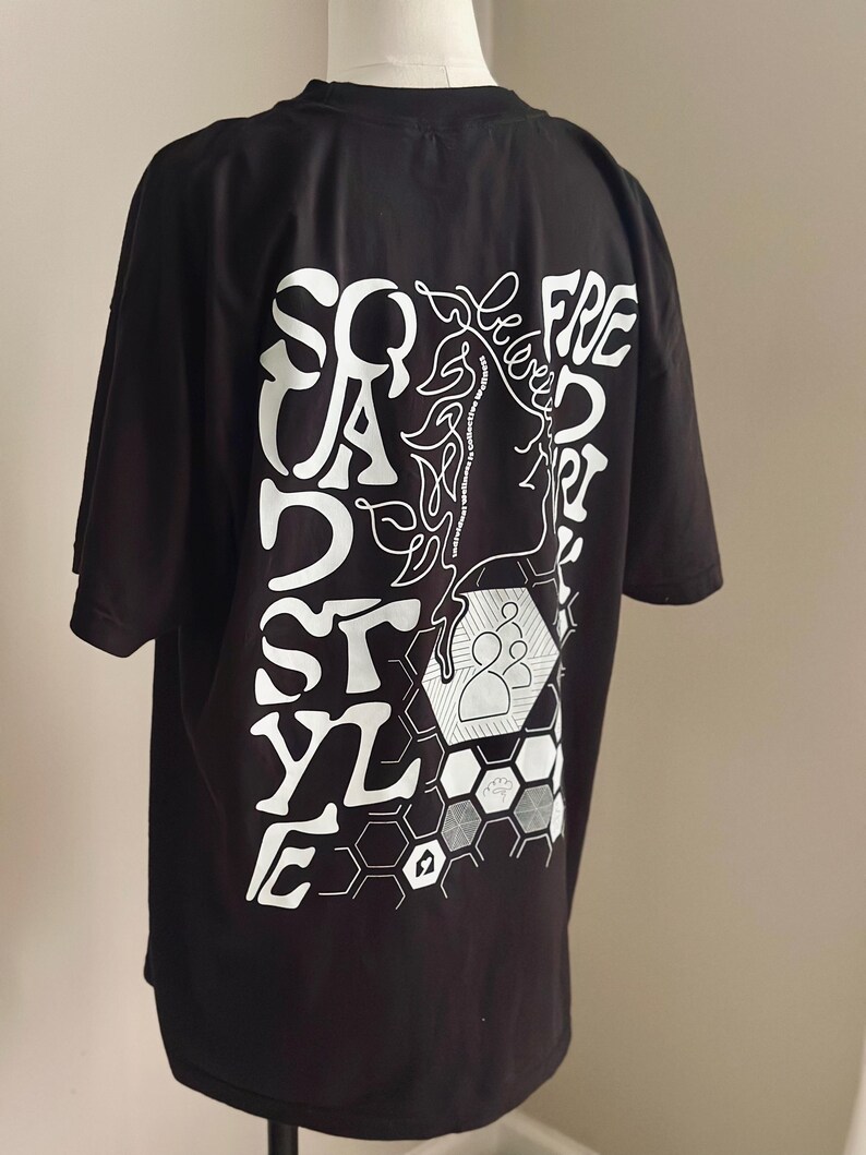 May include: Black t-shirt featuring a white graphic on the back. The design incorporates the words "SQUAD STYLE FREE" in a unique font, a line drawing of a face, and geometric shapes. The t-shirt is displayed on a mannequin.