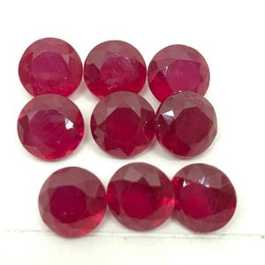 Pode incluir: Nove rubis redondos e facetados são dispostos sobre uma superfície branca. As pedras preciosas exibem uma cor vermelha profunda e uma superfície polida e reflexiva. As gemas são de tamanho e forma semelhantes.