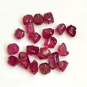 Könnte beinhalten: Eine Sammlung roher, ungeschliffener Rubin-Edelsteine. Die Rubine variieren in Größe und Form und weisen eine tiefrote Farbe auf. Die Edelsteine liegen auf einer weißen Oberfläche und zeigen ihre natürliche, raue Textur.