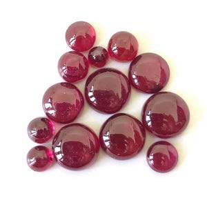 May include: A selection of glossy, round, dark red glass gems of various sizes. The gems are arranged on a white surface, reflecting light. The overall effect is a display of decorative glass stones.