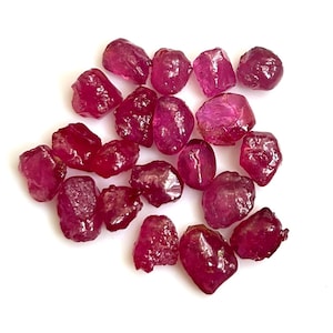 Puede incluir: Un conjunto de gemas de rubí en bruto. Las piedras, de tamaños y formas variadas, presentan un color rojo intenso y una ligera translucidez. Se encuentran dispersas sobre una superficie blanca, resaltando su textura natural y rugosa.