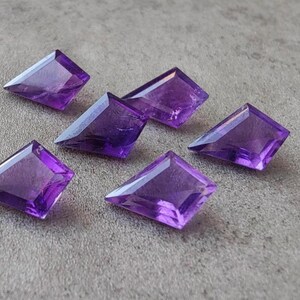 May include: Six faceted, kite-shaped amethyst gemstones. The gemstones are a deep purple color and have a polished, reflective surface. They are arranged on a gray surface.