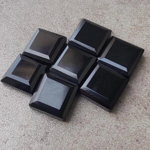 May include: A collection of seven square, black gemstones with beveled edges. The gemstones are arranged in a cluster on a textured, light gray surface. The polished surfaces reflect light, creating highlights.