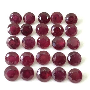 Puede incluir: Veinticuatro rubíes redondos y facetados dispuestos sobre un fondo blanco. Las piedras preciosas son de un color rojo intenso, con facetas visibles que capturan la luz. Los rubíes son de tamaño y forma similares, creando un patrón.