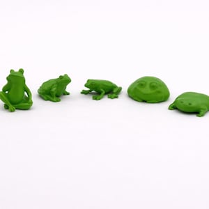 May include: Five small, green frog figurines in various poses are arranged on a white surface. The frogs vary in shape, with some sitting upright and others lying flat. The figurines are a vibrant green colour.