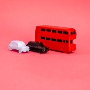 May include: A red double-decker bus, a white car, and a black taxi are arranged on a pink background. The toy vehicles are miniature and appear to be made of plastic. The bus has black windows, and the taxi has black tires.