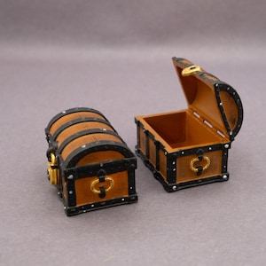 May include: Two miniature treasure chests, one open and one closed. The chests are brown with black trim and gold-coloured hardware. The open chest reveals an empty interior. The closed chest has a curved top and a small lock.