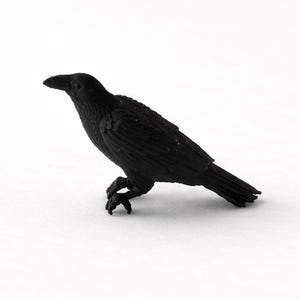 May include: A black raven ornament, set against a white backdrop. The bird is realistically styled, with detailed feathers and a slightly angled pose. The raven is a solid black colour.
