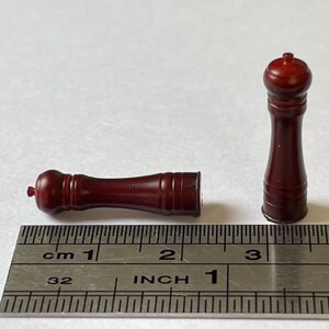 May include: Two wooden salt and pepper shakers, one standing upright and the other lying down, are shown next to a ruler. The ruler is marked in inches and centimeters. The shakers are both a dark reddish brown color.