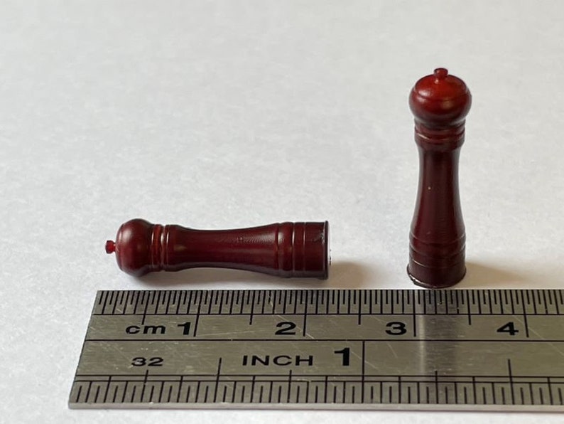 May include: Two wooden salt and pepper shakers, one standing upright and the other lying down, are shown next to a ruler. The ruler is marked in inches and centimeters. The shakers are both a dark reddish brown color.