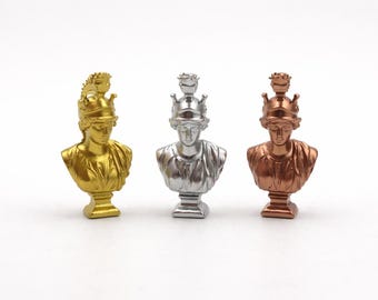 May include: Three metallic busts of a classical figure, each with a detailed helmet and draped garment. The busts are in gold, silver, and copper colors, standing on a white background. The statues are decorative.