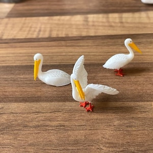May include: Three white pelican figurines with orange beaks and feet. The pelicans are posed in different positions, with one standing upright, one with its head turned to the side, and one with its wings spread out.