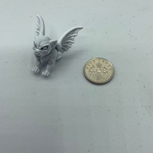 May include: A small, white, winged gargoyle figurine with a detailed face. The figurine is about the size of a US quarter.