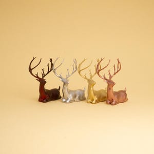 May include: Four reindeer figurines in different colours: red, silver, gold, and copper. The reindeer are all in a sitting position with their heads turned to the side.