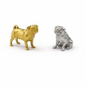 May include: Two pug dog ornaments, one gold and one silver, are set against a white backdrop. The gold pug is standing, while the silver pug is seated. Both ornaments have detailed features.