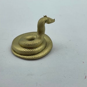 May include: A gold-coloured snake figurine curled up in a coiled position. The snake has a textured surface and a slightly raised head.