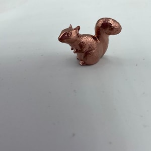 May include: A small, copper-colored squirrel figurine. The squirrel is sitting on its haunches with its tail curled up behind it.