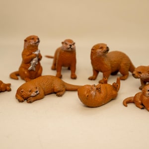 May include: A collection of eight brown otter figurines in various poses. One otter stands holding a silver fish, while others are depicted lying down, sitting, or resting on their backs. The figures feature textured fur details and realistic facial expressions.