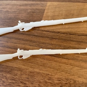 May include: Two white plastic toy rifles are displayed on a wooden surface. The rifles have detailed features, including a trigger, stock, and barrel. The toy guns are likely for play or display.