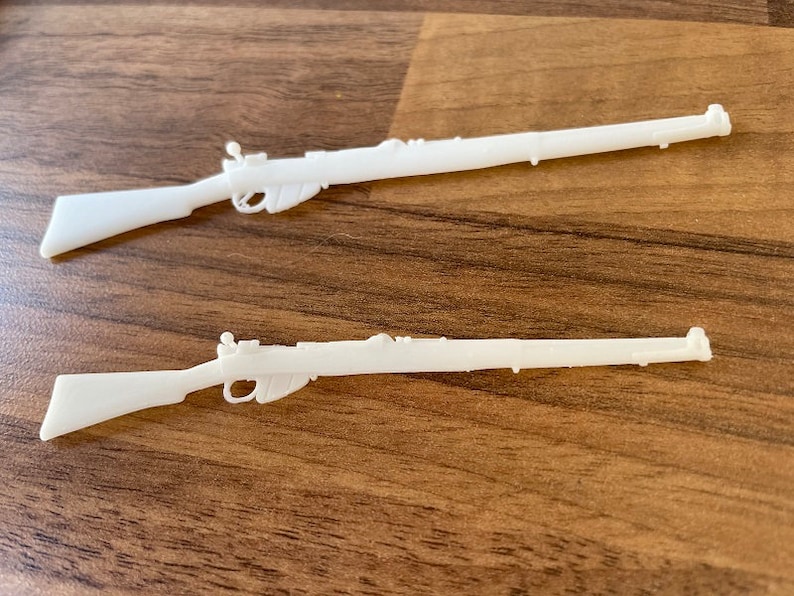 May include: Two white plastic toy rifles are displayed on a wooden surface. The rifles have detailed features, including a trigger, stock, and barrel. The toy guns are likely for play or display.