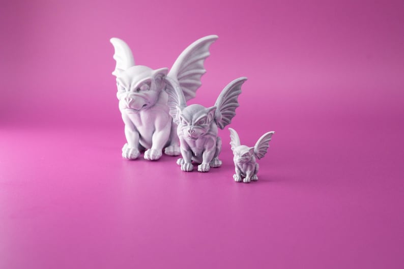 May include: Three white gargoyle figurines of varying sizes, with wings and a fierce expression, on a pink background.