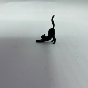 May include: A small, solid black figurine of a cat in a stretching pose. The cat is depicted with its front paws extended forward, its back arched upward, and its tail held straight up in the air, capturing a classic feline stretch silhouette.
