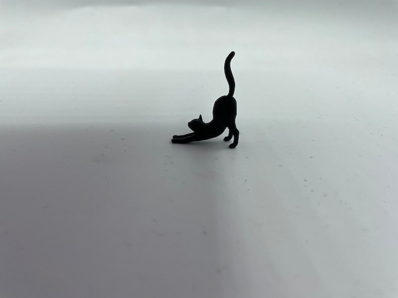 May include: A small, solid black figurine of a cat in a stretching pose. The cat is depicted with its front paws extended forward, its back arched upward, and its tail held straight up in the air, capturing a classic feline stretch silhouette.