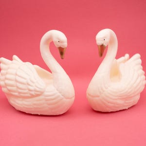May include: Two white swan-shaped figurines with textured feather details and gold-colored beaks. The swans are positioned facing each other, each featuring a curved neck and a hollowed-out center area, presented against a solid pink background.