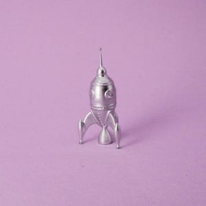May include: A silver toy rocket ship with a pointed nose and two small wings on the side. The rocket is standing on a light purple surface.