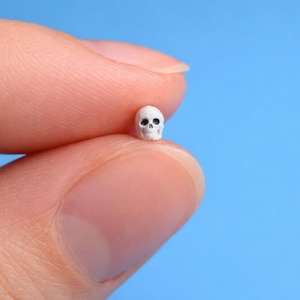 May include: A miniature white skull with black eye sockets is held between two fingers against a bright blue backdrop. The tiny skull is a detailed replica, showcasing the intricate structure of a human skull.