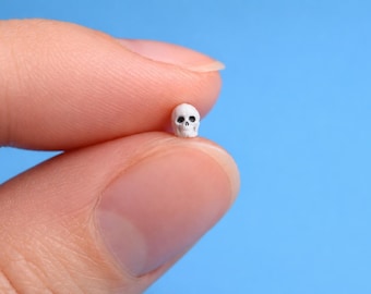 May include: A tiny, white skull with black eye sockets is held between two fingers against a bright blue background. The miniature skull is a detailed replica, showcasing the intricate structure of a human skull.