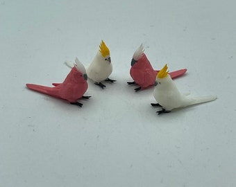 May include: Four miniature bird figurines, two white with yellow crests and two pink with white crests. The figurines are arranged in a circle on a white surface.