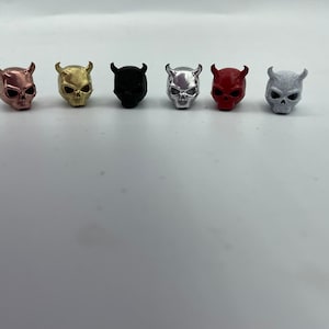 May include: Six small devil skull figurines in a row. The skulls are in various metallic colors: copper, gold, black, silver, red, and gray. Each skull has horns and a detailed facial structure.