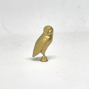 May include: A gold-colored owl figurine with detailed features. The owl is standing on one leg with its wings folded.