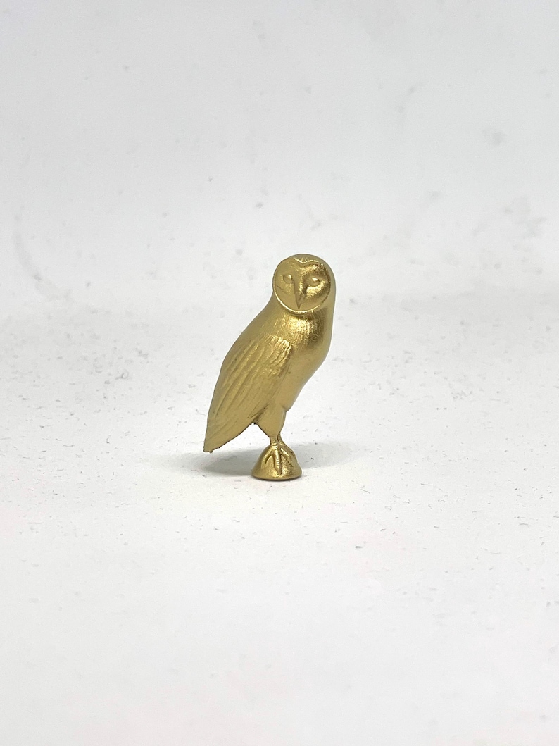 May include: A gold-colored owl figurine with detailed features. The owl is standing on one leg with its wings folded.
