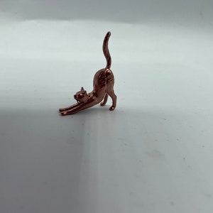 May include: A small, metallic copper-colored figurine of a cat in a stretching pose. The cat is depicted with its front paws extended forward, its back arched upward, and its tail held straight up in the air, capturing a dynamic movement.