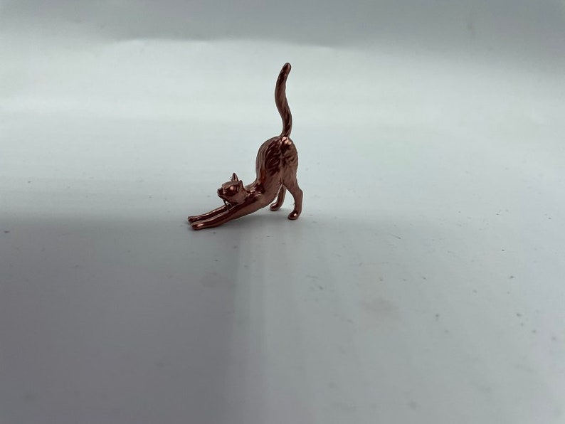 May include: A small, metallic copper-colored figurine of a cat in a stretching pose. The cat is depicted with its front paws extended forward, its back arched upward, and its tail held straight up in the air, capturing a dynamic movement.