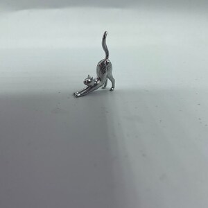 May include: A small, silver-colored metal figurine of a cat in a stretching pose. The cat is depicted with its front paws extended forward, its back arched upward, and its tail pointing straight toward the sky, capturing a classic feline stretch.