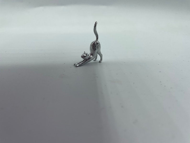 May include: A small, silver-colored metal figurine of a cat in a stretching pose. The cat is depicted with its front paws extended forward, its back arched upward, and its tail pointing straight toward the sky, capturing a classic feline stretch.