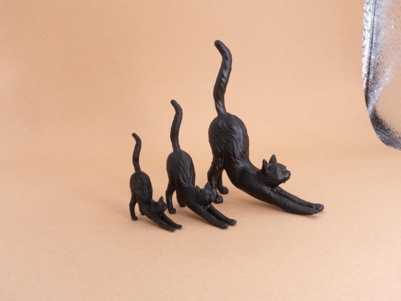May include: Three black cat figurines in graduated sizes, each depicted in a stretching pose with front legs extended forward, arched backs, and tails held upright. The figures feature textured surfaces representing fur and are arranged in a row.