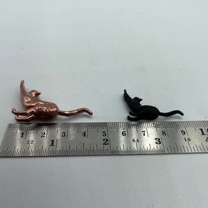 May include: A metallic copper-colored cat figurine and a smaller matte black cat figurine positioned next to a stainless steel ruler. The copper cat measures approximately 1.5 inches in length, while the black cat measures about 1 inch long.