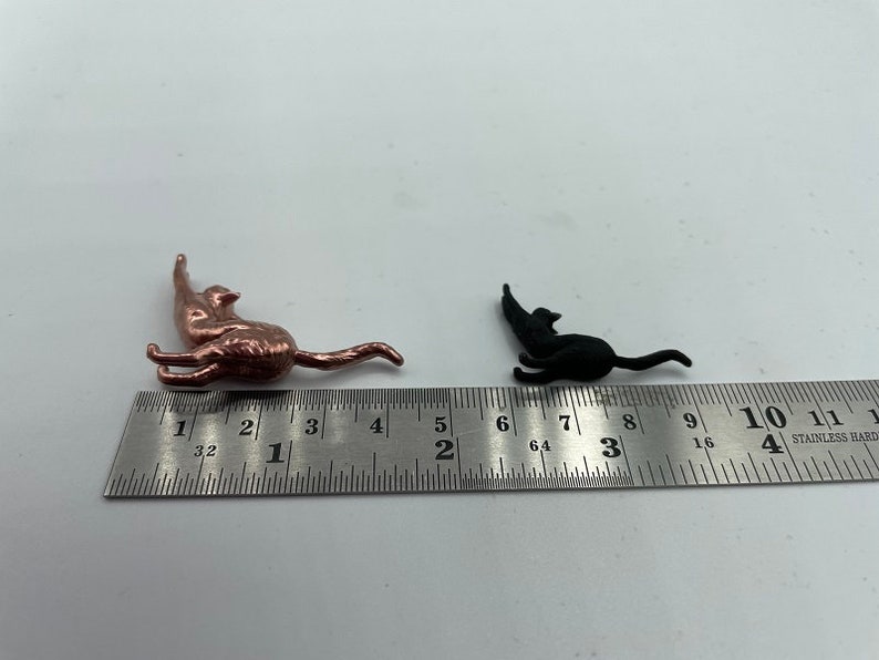May include: A metallic copper-colored cat figurine and a smaller matte black cat figurine positioned next to a stainless steel ruler. The copper cat measures approximately 1.5 inches in length, while the black cat measures about 1 inch long.