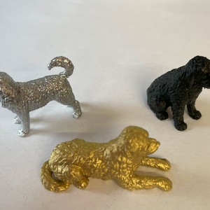 Peut inclure: Trois petites figurines de chiens, une argentée, une dorée et une noire. Les figurines argentée et dorée sont debout, tandis que la figurine noire est assise.