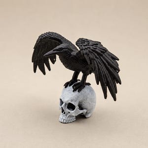 以下が含まれることがあります： 翼を広げた黒いカラスのフィギュアが、リアルな人間の頭蓋骨の上に止まっています。カラスには詳細な羽の質感と鋭い嘴があり、頭蓋骨はオフホワイトで、縫合線と深い眼窩がはっきりと見えます。