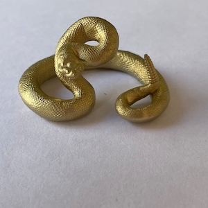 May include: A gold-coloured, 3D printed snake figurine coiled in a circle. The snake has a textured surface and a realistic appearance.