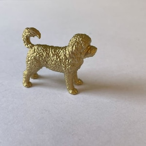 May include: A small, gold-coloured dog figurine with a curly coat and a curled tail. The dog is standing on all fours, with its head turned slightly to the right. The figurine is made of a shiny, metallic material.