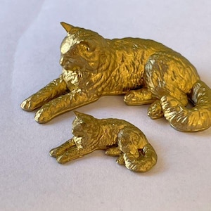 May include: Two gold-painted cat figurines, one larger than the other, are posed in a sitting position. The larger cat has its tail curled around its body, while the smaller cat is lying down with its head turned to the side.