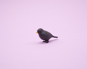 May include: A small black bird figurine with a yellow beak, standing on a light purple background.